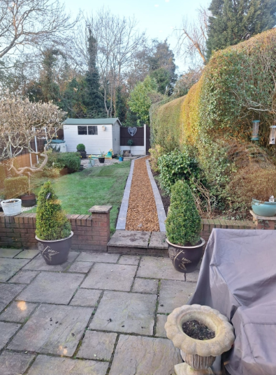 Backyard garden with stone patio, lawn, gravel path, hedges, and a small shed in the distance