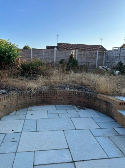 Stone patio with curved brick wall, overgrown dry grass, and backyard fence under a clear sky