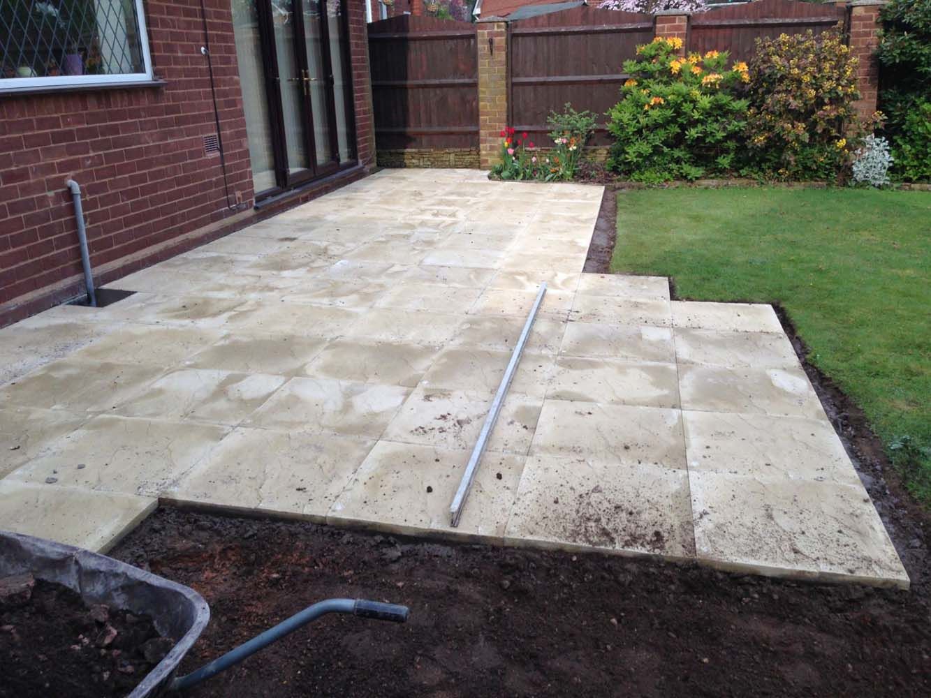 Paved backyard patio beside a lawn, with a wooden fence and brick house wall.