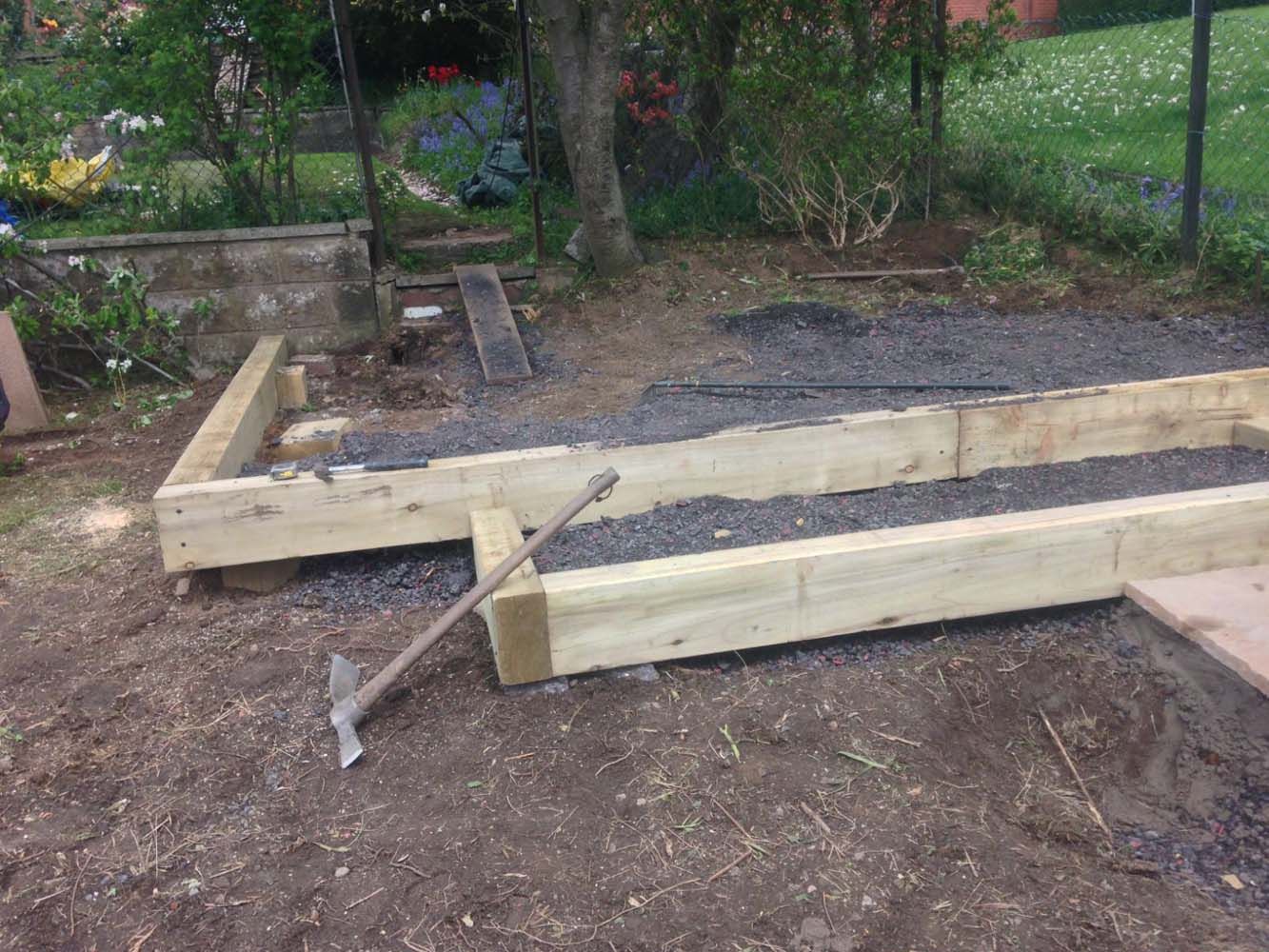 Wooden garden frame under construction with boards, braces, and gravel beside a fence and trees.