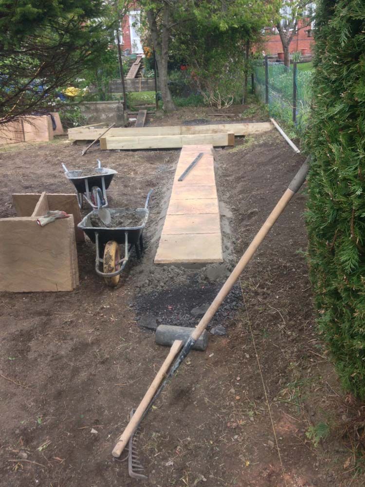 Garden path under construction with shovel, wheelbarrow, and wooden boards along a dirt trench.
