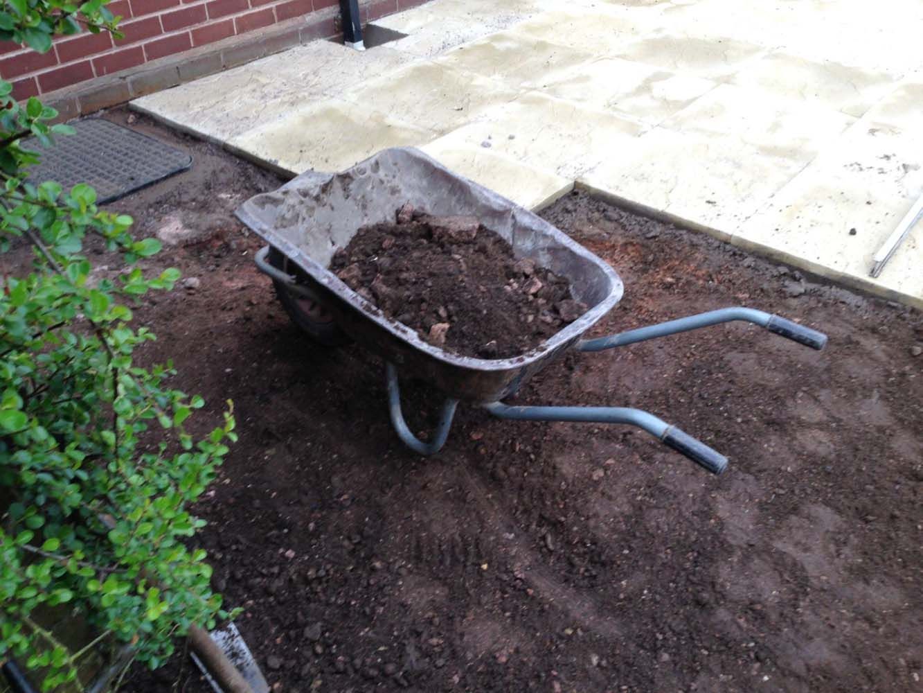 Wheelbarrow filled with soil beside a garden bed and patio pavers