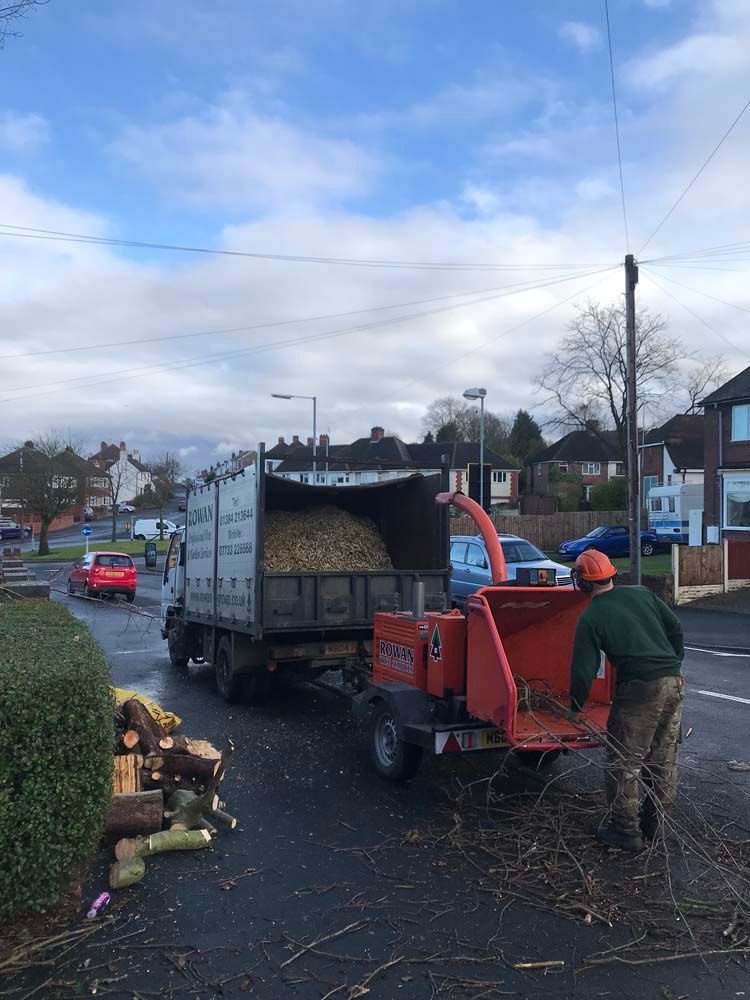 Worker feeding wood chipper from truck on suburban street, with another person nearby and piles of branches.