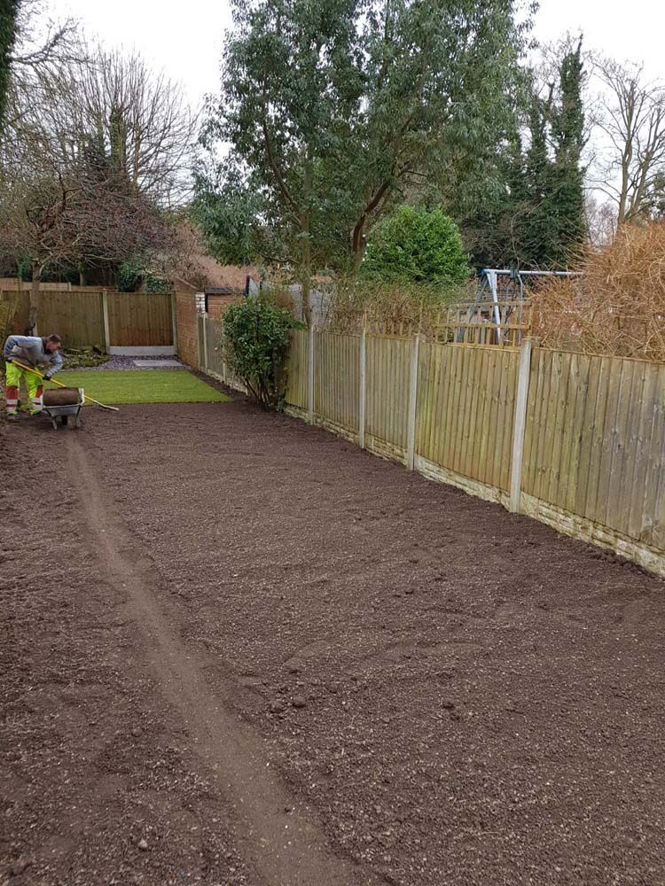 Freshly tilled garden soil beside a fence, with a small tiller and trees in the background