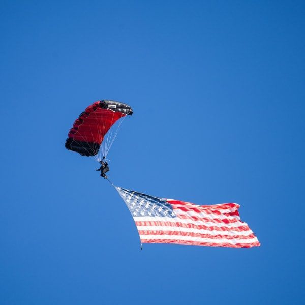 Parachutist with red parachute carrying the American flag against a clear blue sky.