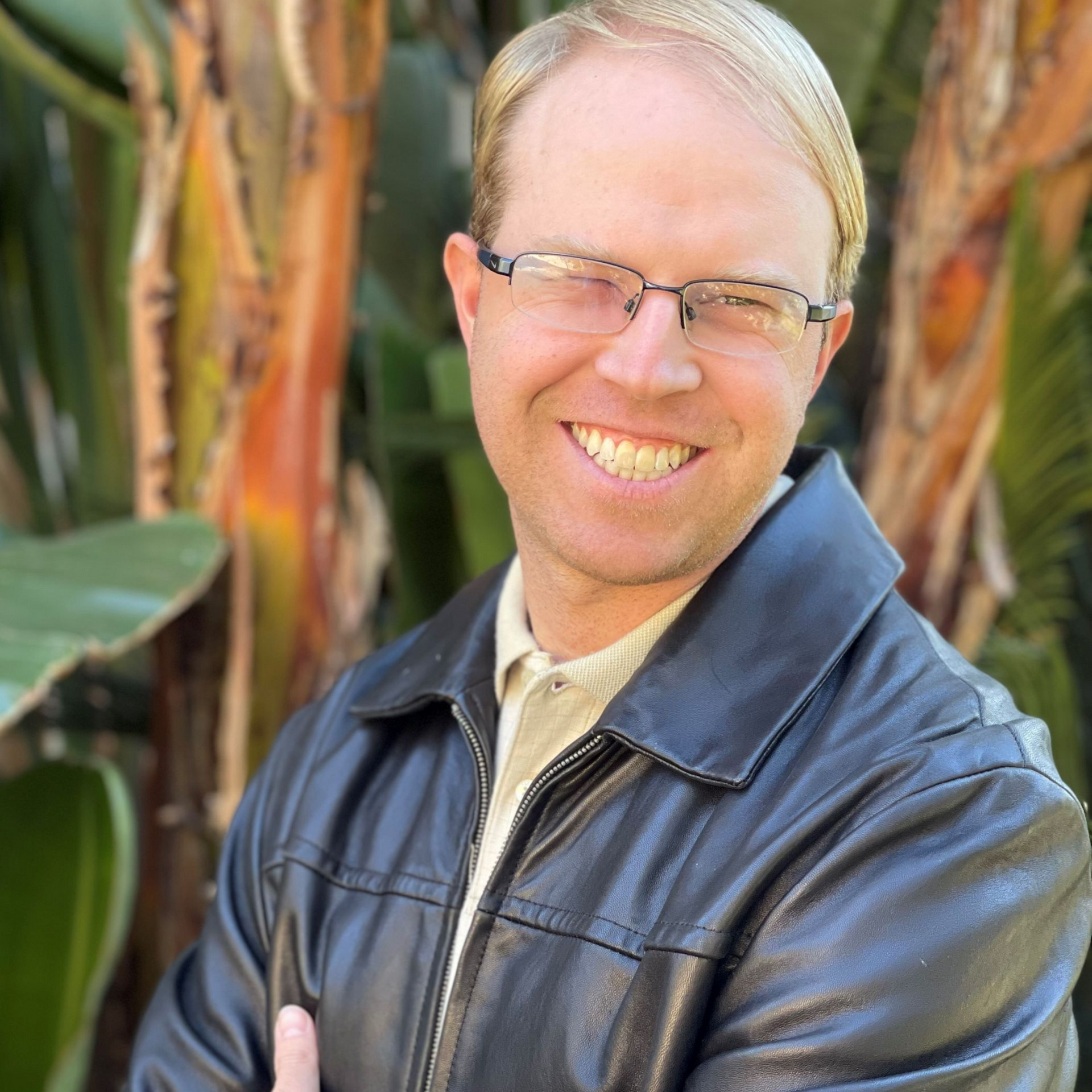 Man with blond hair wearing glasses and a light blue polo shirt smiles outside.