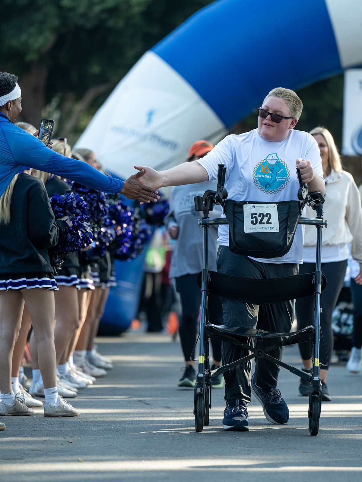 Person with walker shakes hands at a race start, cheered on by a line of cheerleaders and an archway.