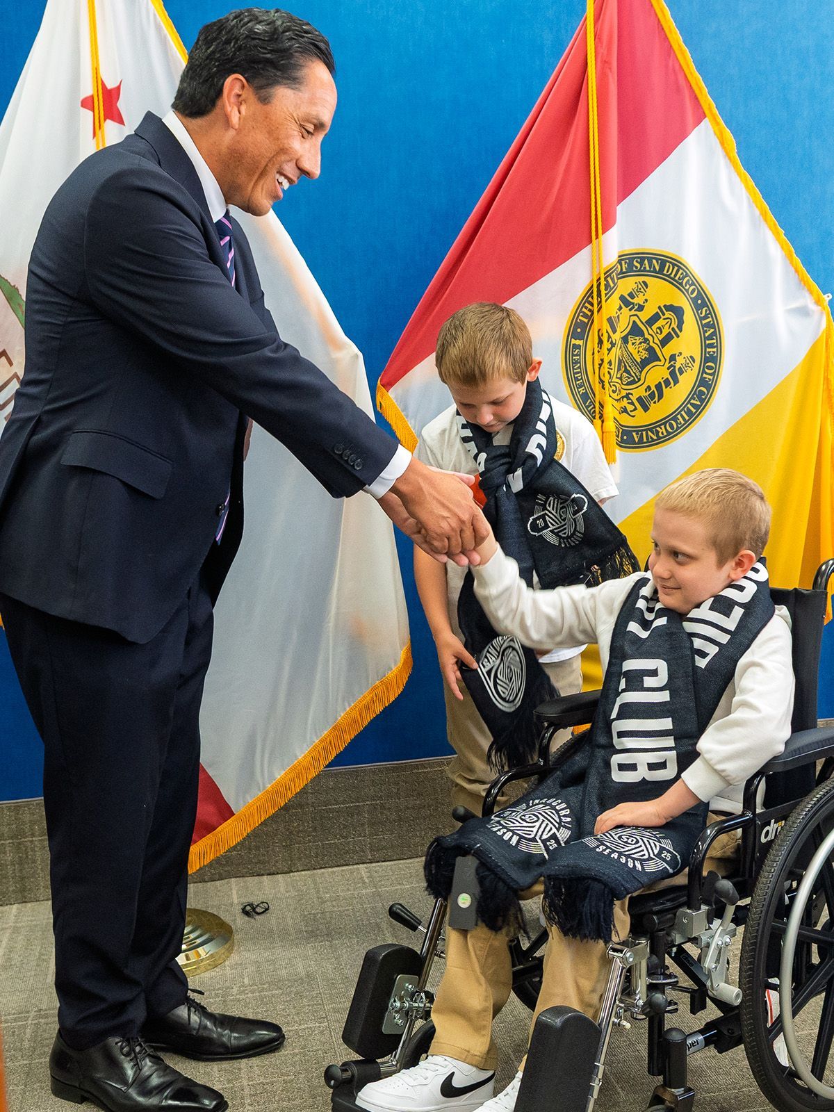 Man shakes hands with child in a wheelchair, children with scarves stand by flags.