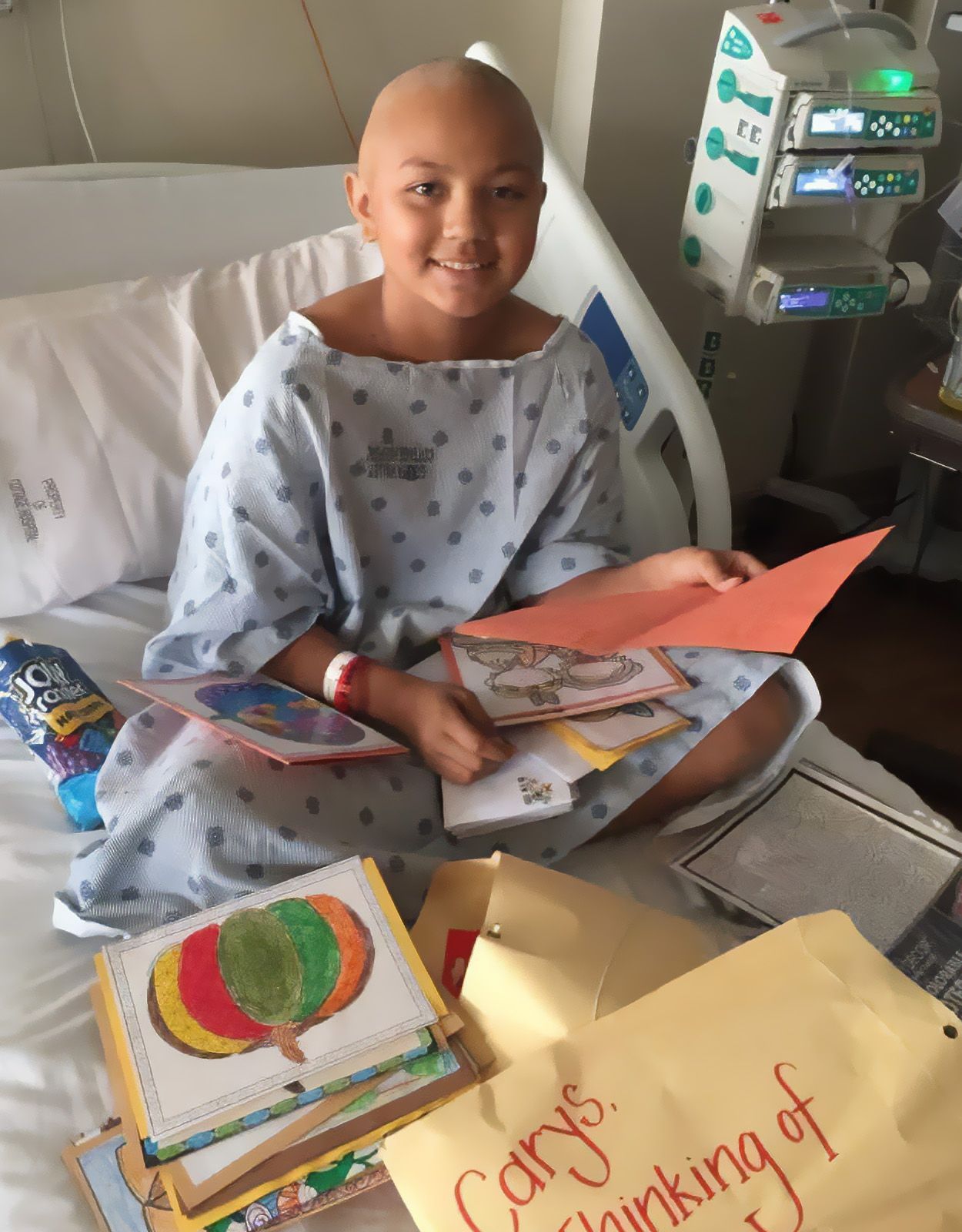 Child in hospital gown, bald head, smiles, holding get-well cards in a hospital bed.