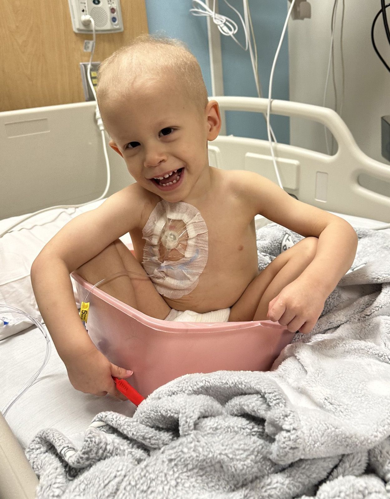 Smiling child with a surgical dressing on his chest, sitting in a pink basin in a hospital bed.