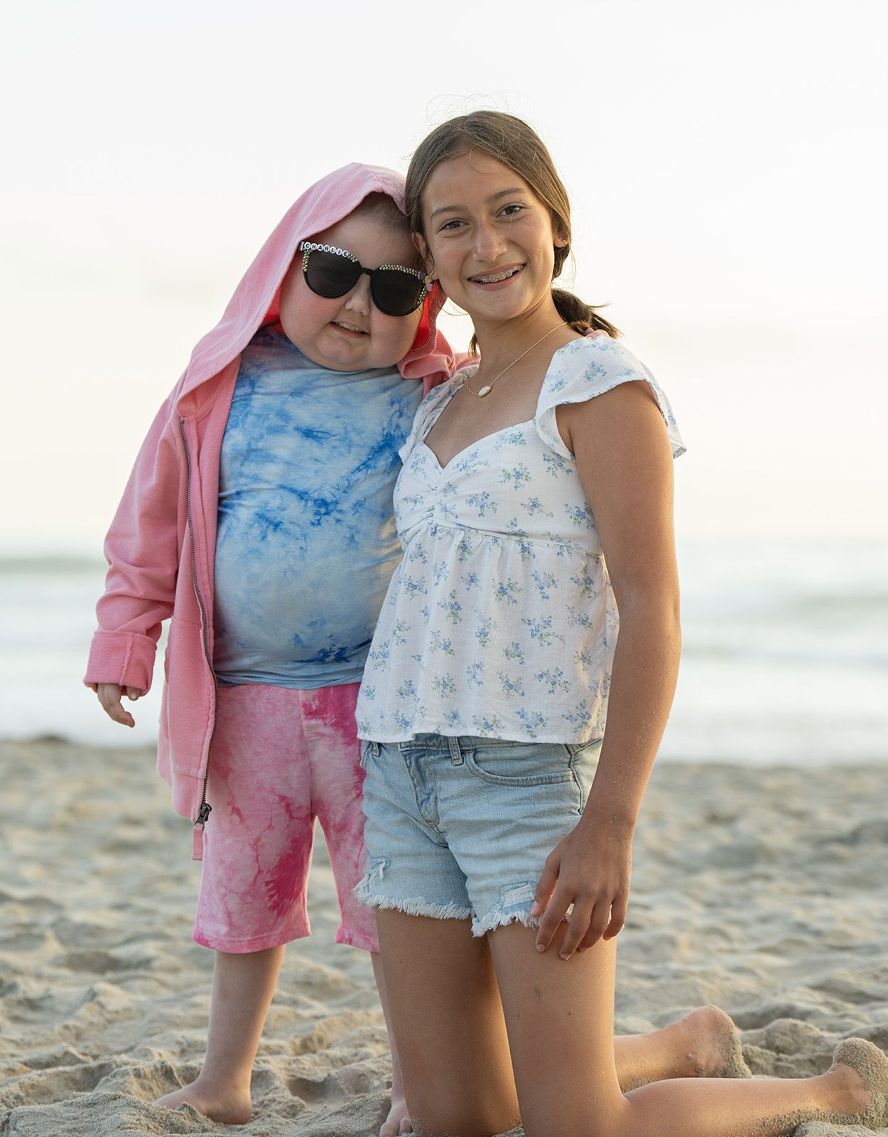 Two girls on a beach: one in sunglasses and pink outfit, other in denim shorts and white top.