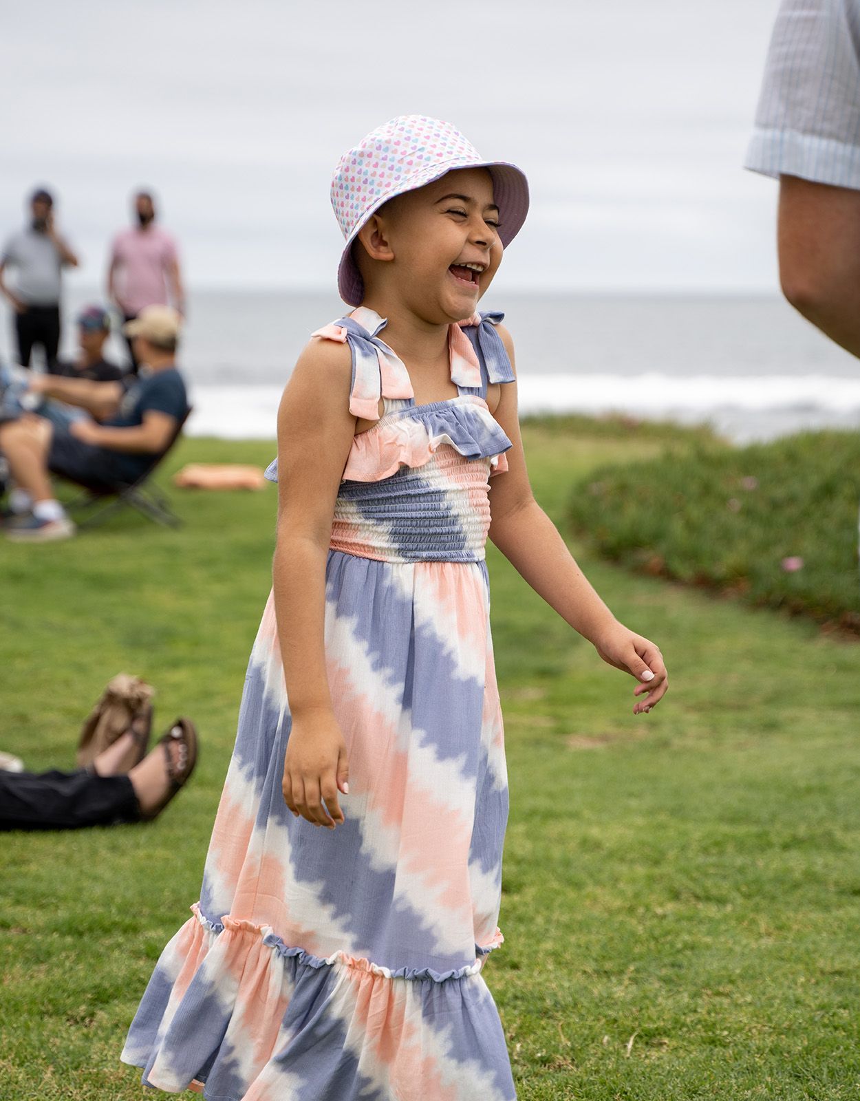 Girl in sun hat and tie-dye dress laughs on green grass by ocean.