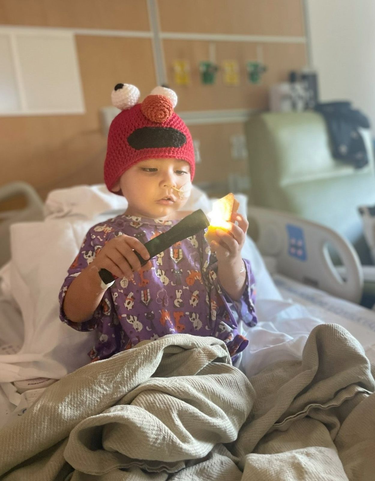 Child in Elmo hat, holding a flashlight in a hospital bed.