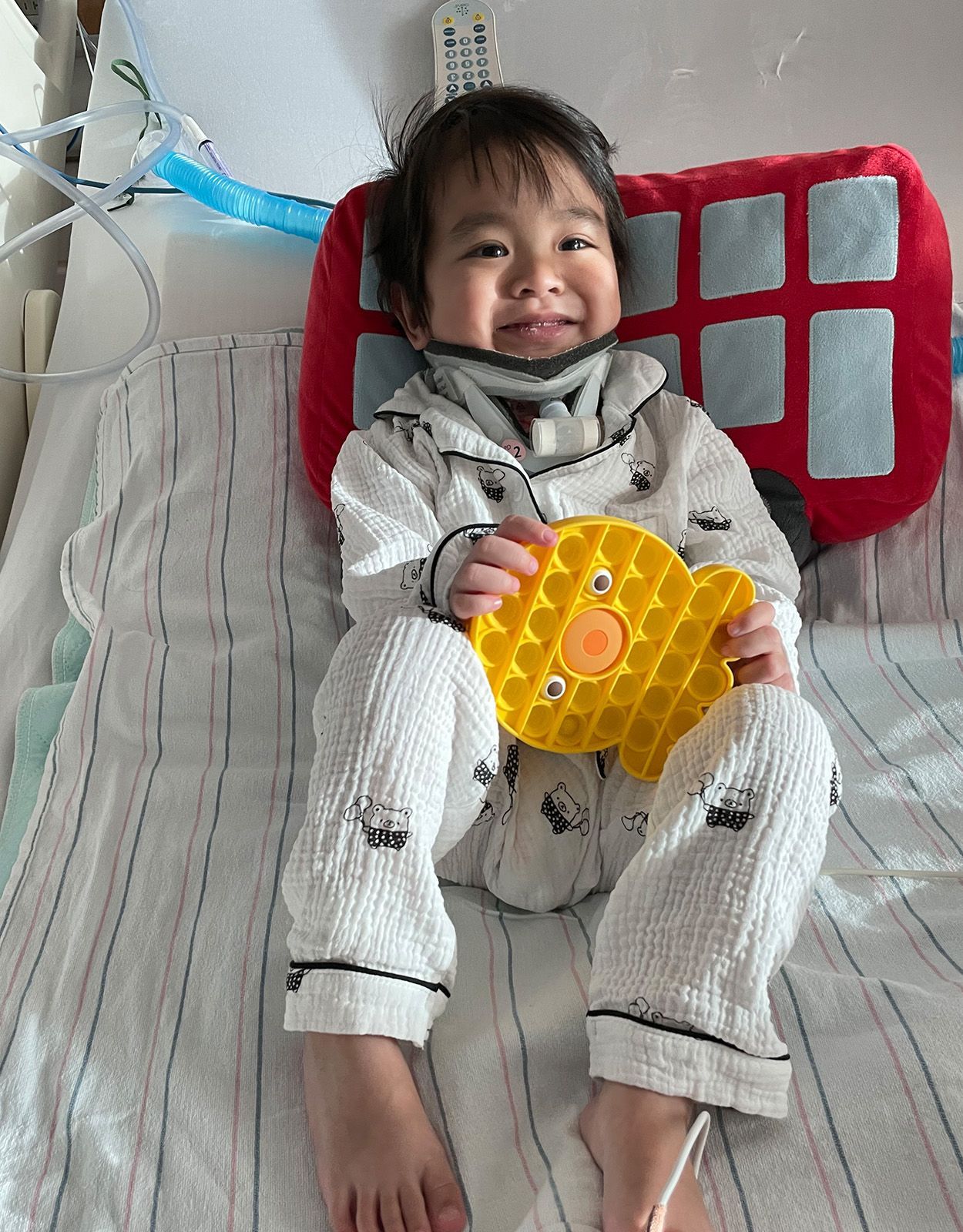 Boy in pajamas holding a yellow toy, sitting up in a hospital bed with a red pillow behind him.