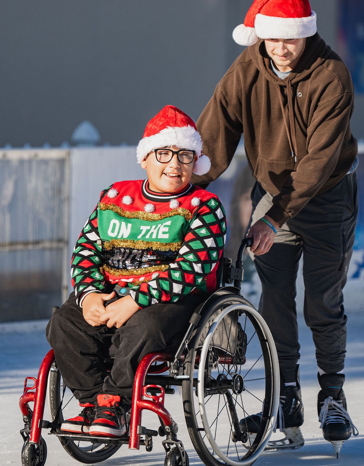 Boy in wheelchair on ice rink wearing Christmas sweater and Santa hat, being pushed by another boy on skates.