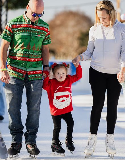 Family ice skating: father in green shirt, mother, and child in red sweater holding hands, smiling.