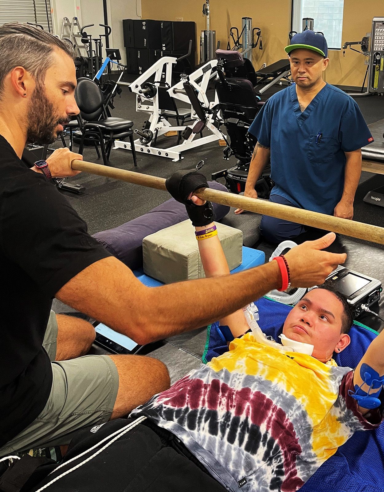 Man helps another using a stick. Person is in chair. Trainer watches in gym.