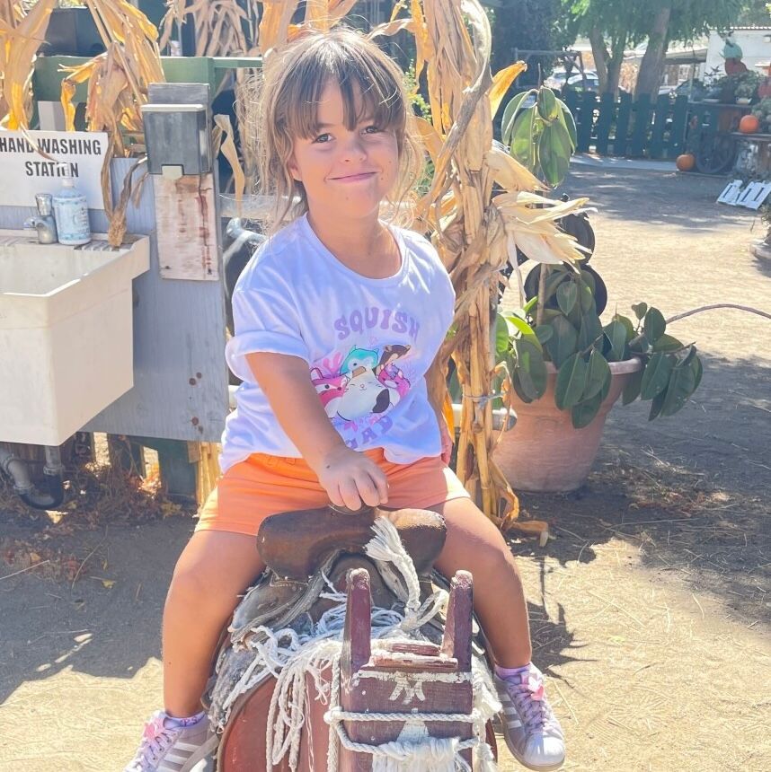 A person sits on a decorative wooden horse at an outdoor pumpkin patch, wearing a white shirt and orange shorts.