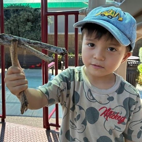 A child in a blue construction hat and Mickey Mouse shirt holds a Y-shaped stick outdoors.