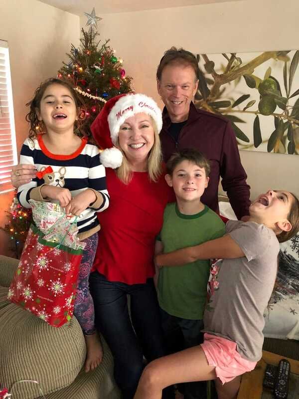 Family of five smiles near a Christmas tree. Woman in a Santa hat, red shirt. Kids hold stocking, laugh.
