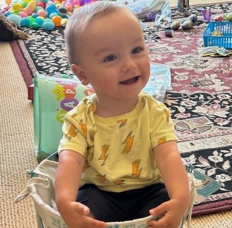 A smiling child wearing a yellow shirt with lightning bolts sits inside a light-colored basket on a patterned rug.