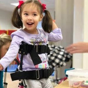 Young girl smiles while wearing a mobility aid. Indoors, she has two pink bows in her hair.