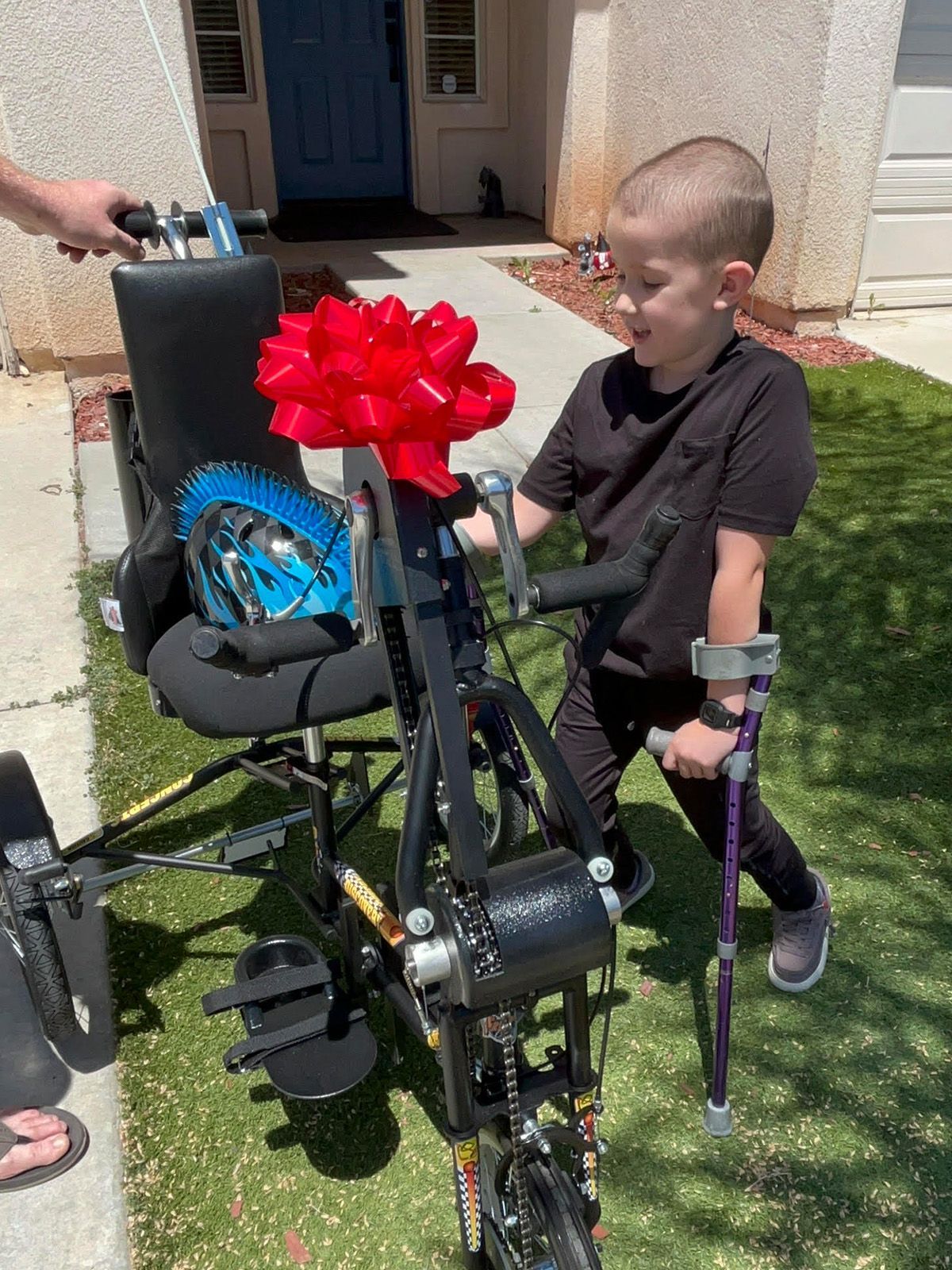 Boy with crutches smiles at his new adaptive bike, decorated with a red bow, outside.