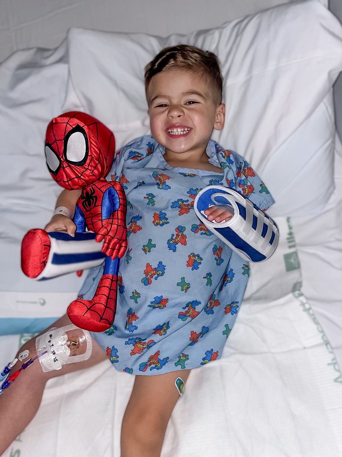 Smiling young boy in hospital bed, holding Spider-Man toy, wearing a hospital gown and arm brace.