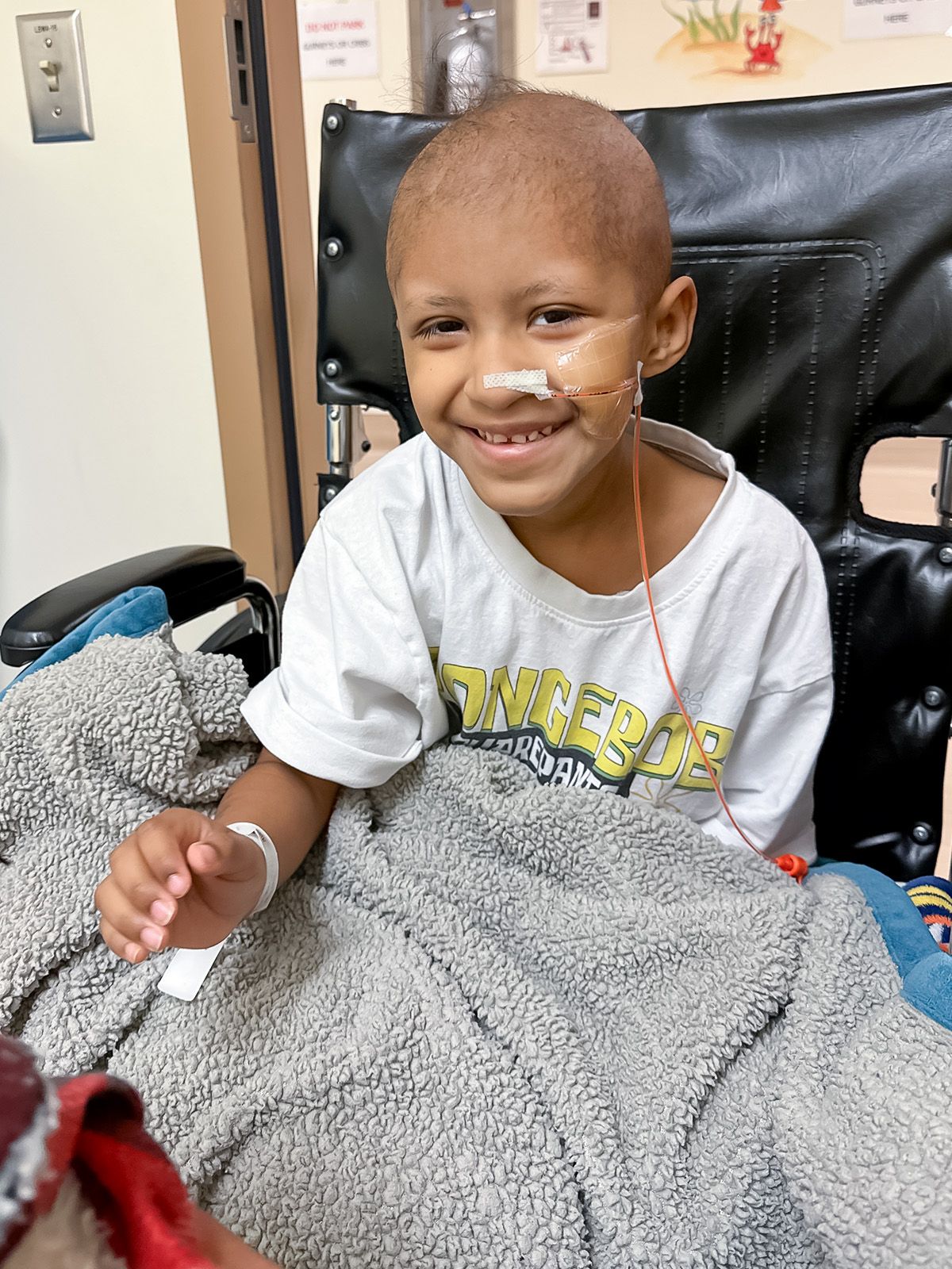 Young child with a shaved head smiles while sitting in a wheelchair. They have a feeding tube and a hospital setting.