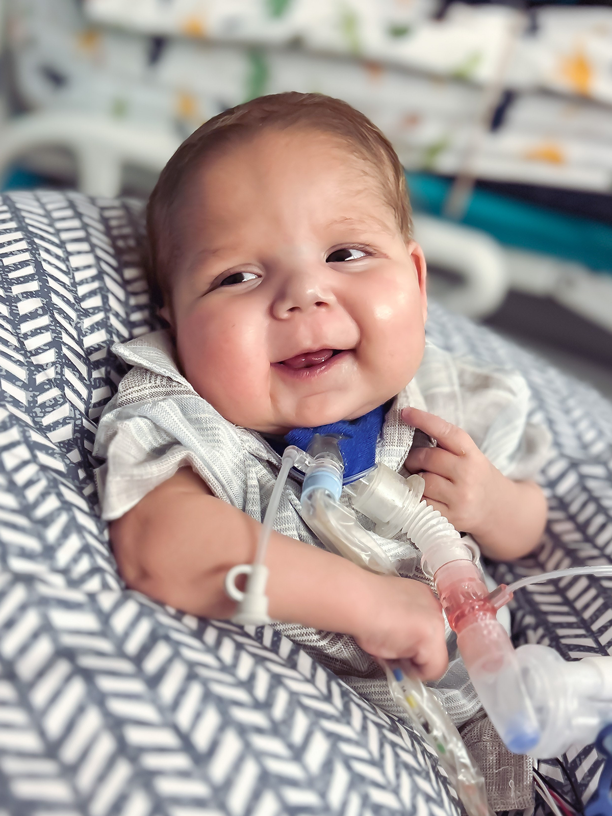 Smiling baby with breathing tube, in hospital setting, resting on a patterned pillow.