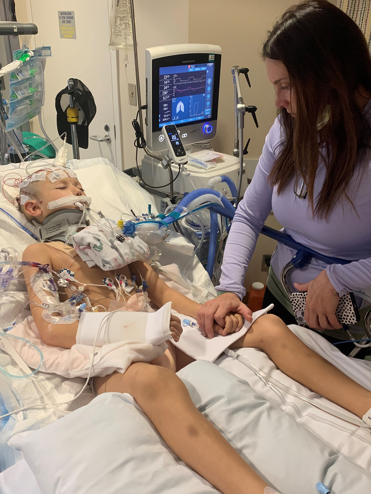Woman holds child's hand in hospital bed, child connected to life support.