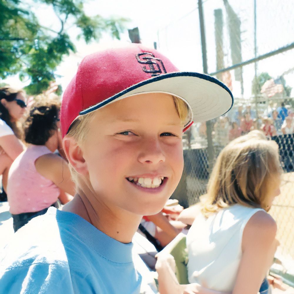 Boy in red baseball cap smiles at a game, with blurred crowd and fence in background.
