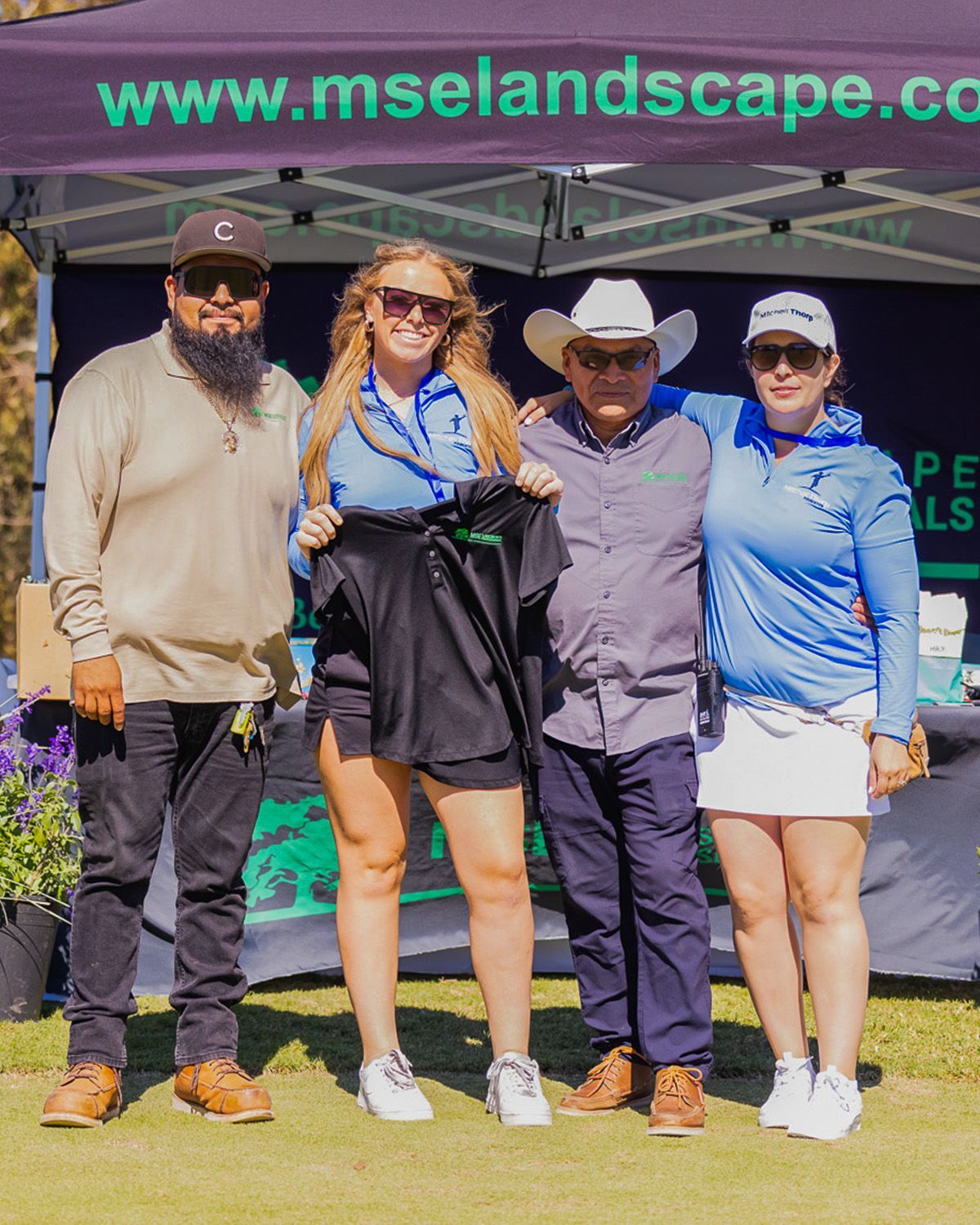 Four people pose for a photo in front of a company tent at a golf course, with one person holding up a black golf shirt.