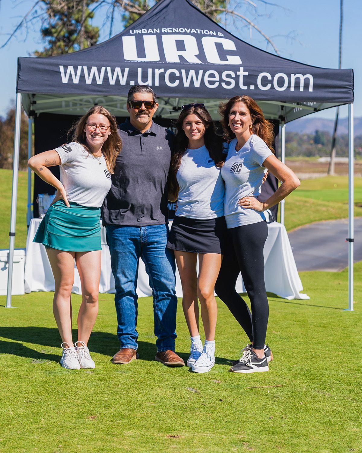 Four people stand smiling in front of a URC West branded tent on a sunny golf course.