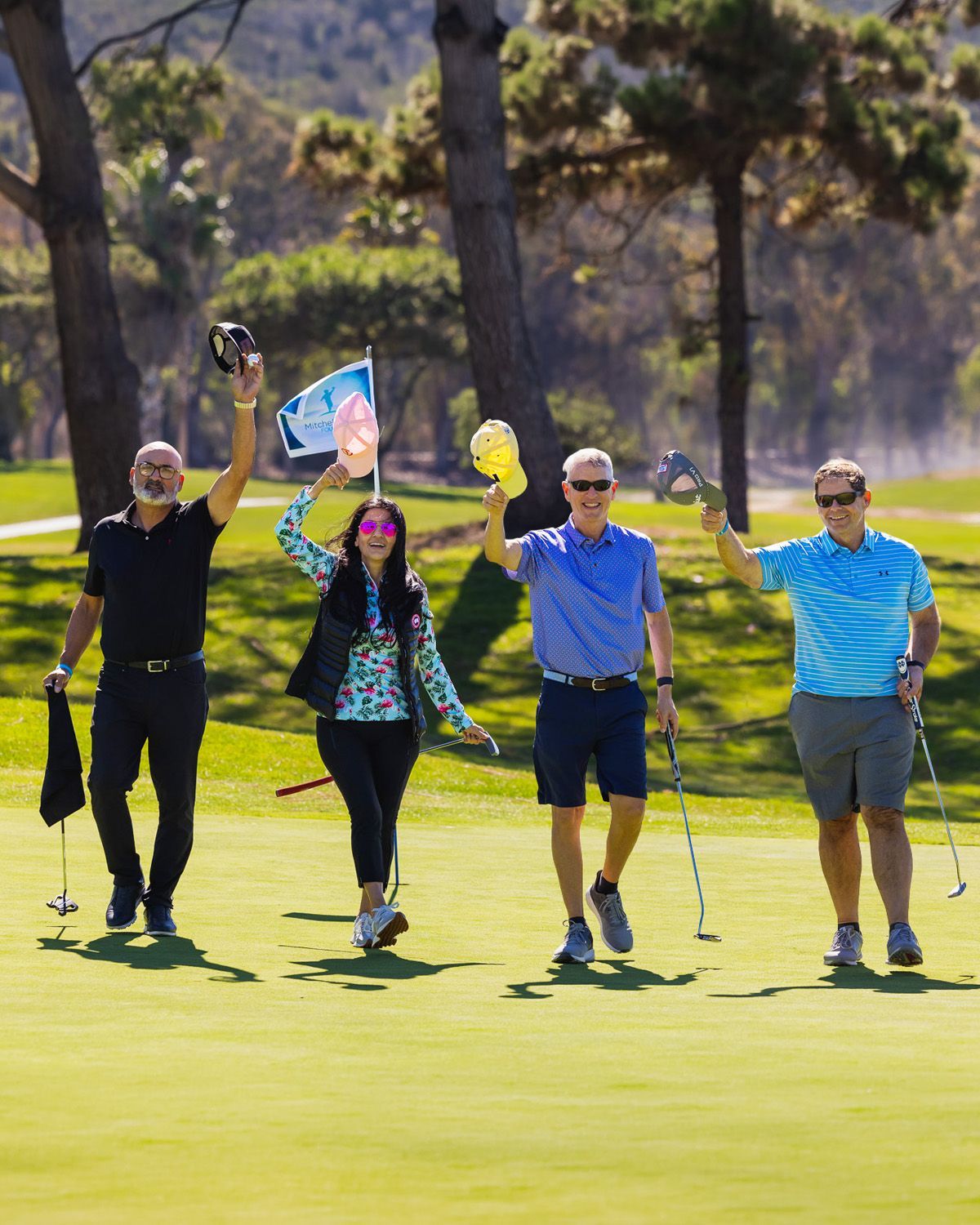 Four people walk on a sunny golf course, smiling and holding golf clubs, flags, and balls while walking toward the camera.