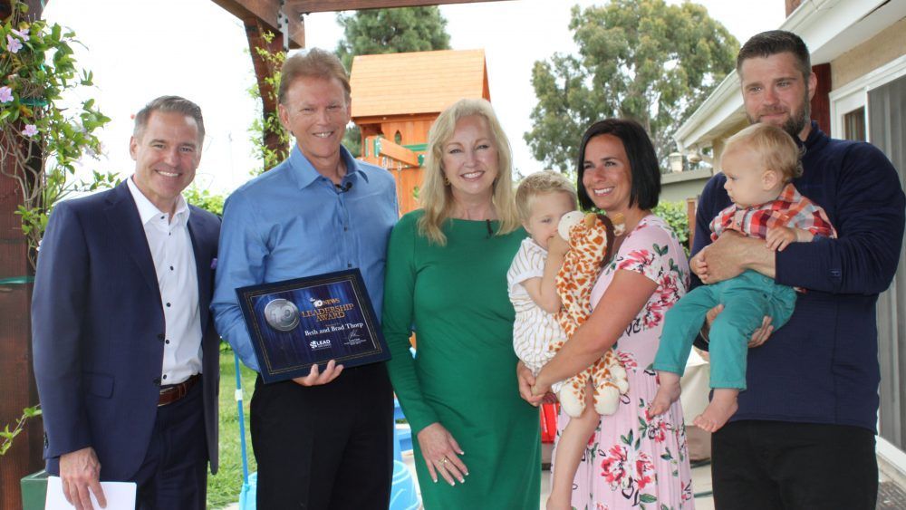 Group of people posing with a plaque. Two women hold babies. Outdoors, sunny setting.
