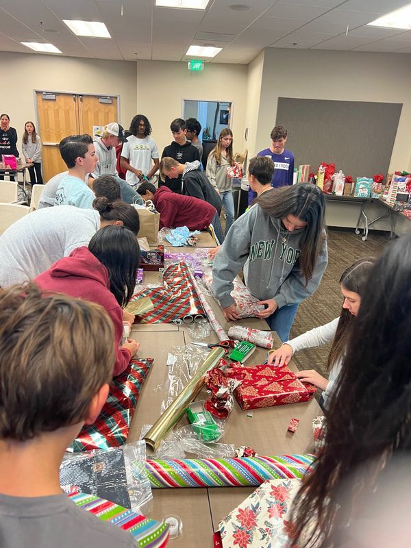 A group of people wrapping gifts at a table. Wrapping paper and presents are on the table.