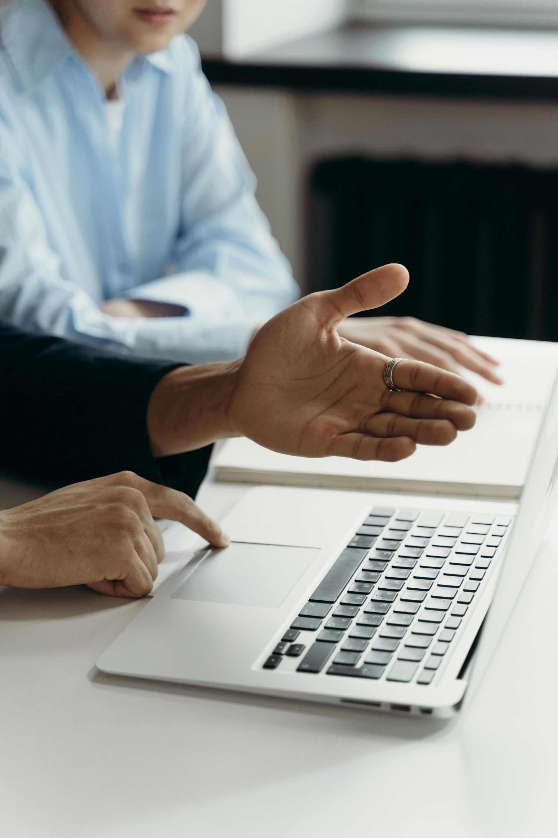 Two people collaborate at a table as one person points to a laptop screen while the other gestures with an open hand.