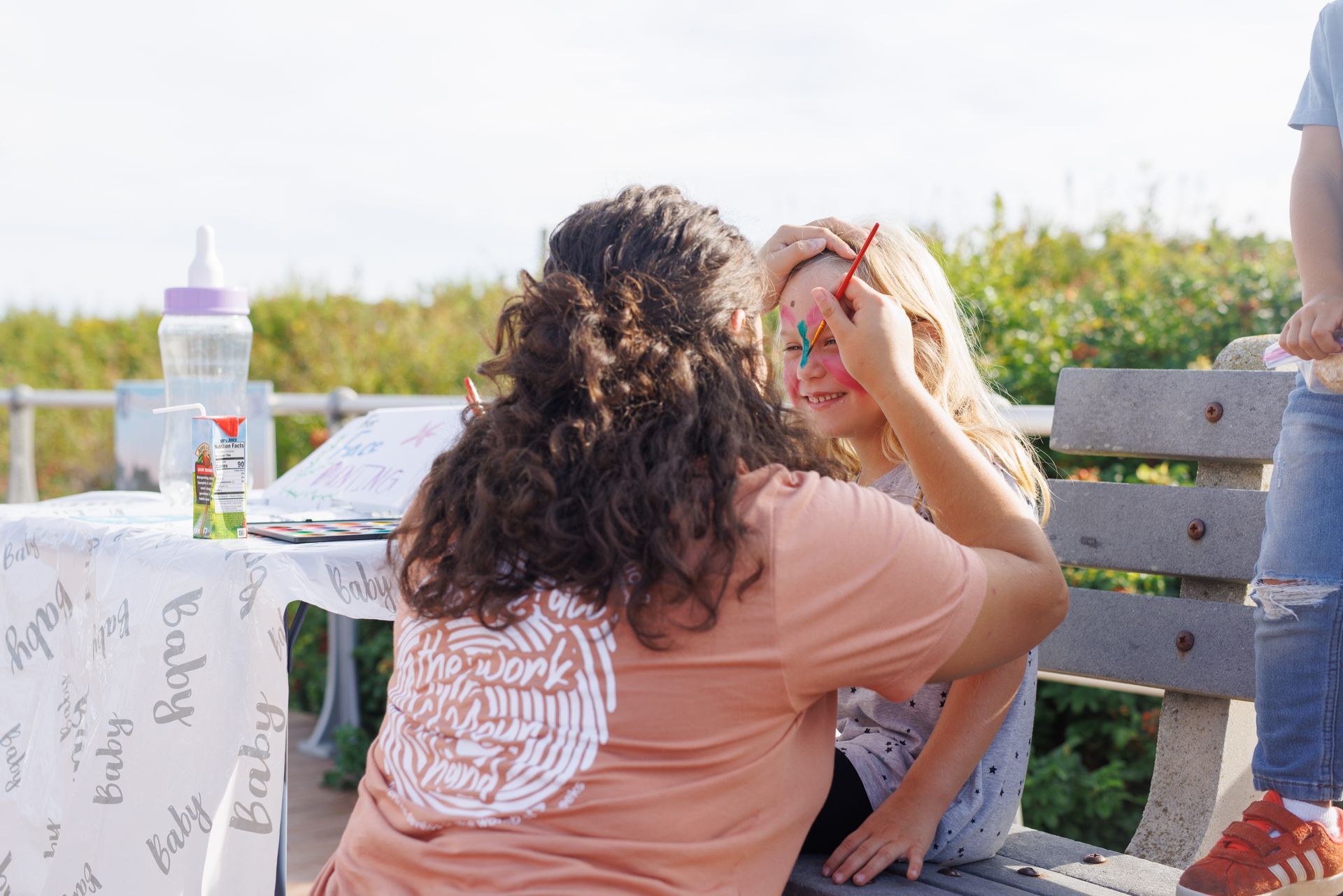 Woman paints a butterfly face on a young girl seated on a bench outdoors. A table with supplies is in the background.