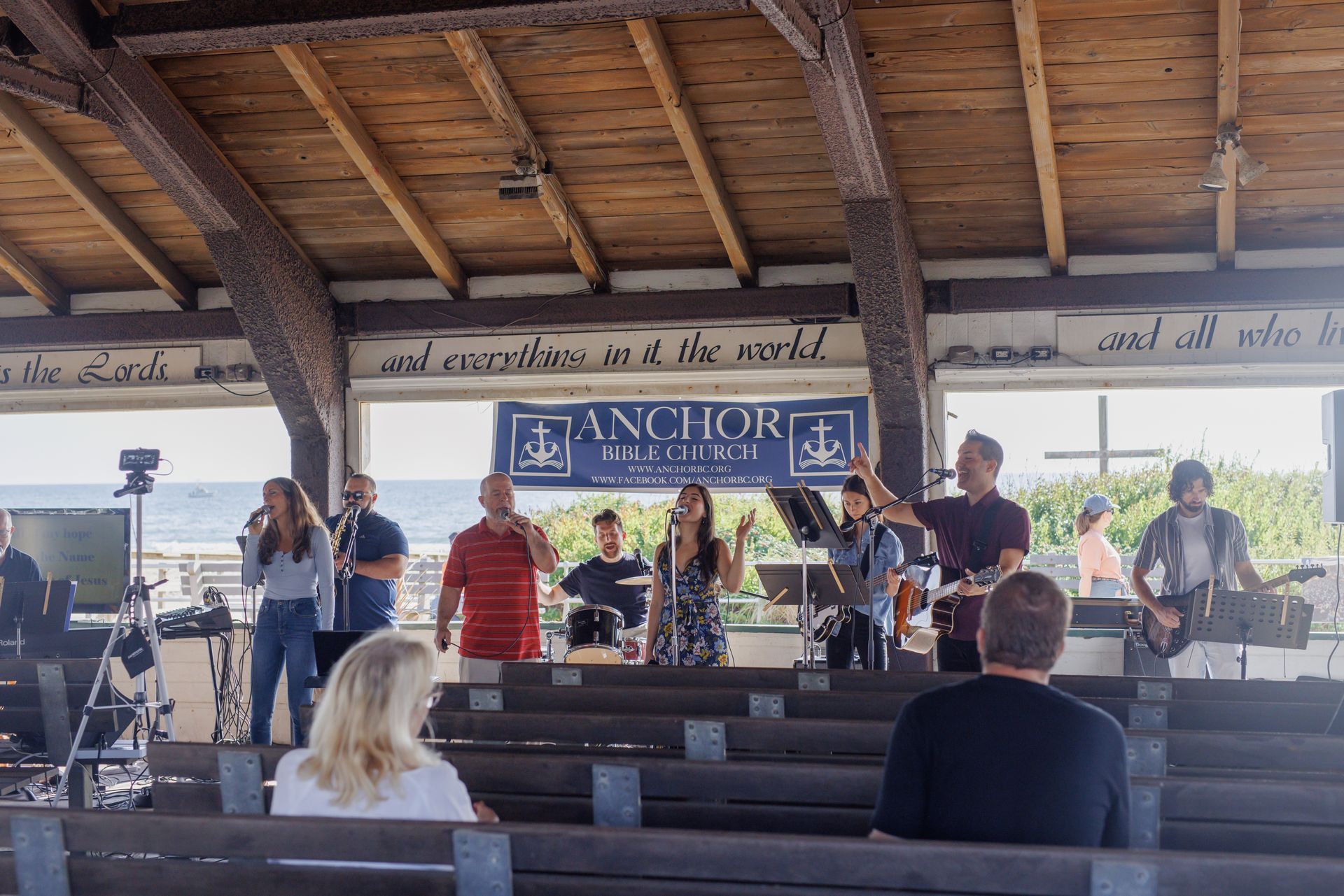 Band performing worship music outdoors under a wooden shelter. People raise hands. Blue and white banner.