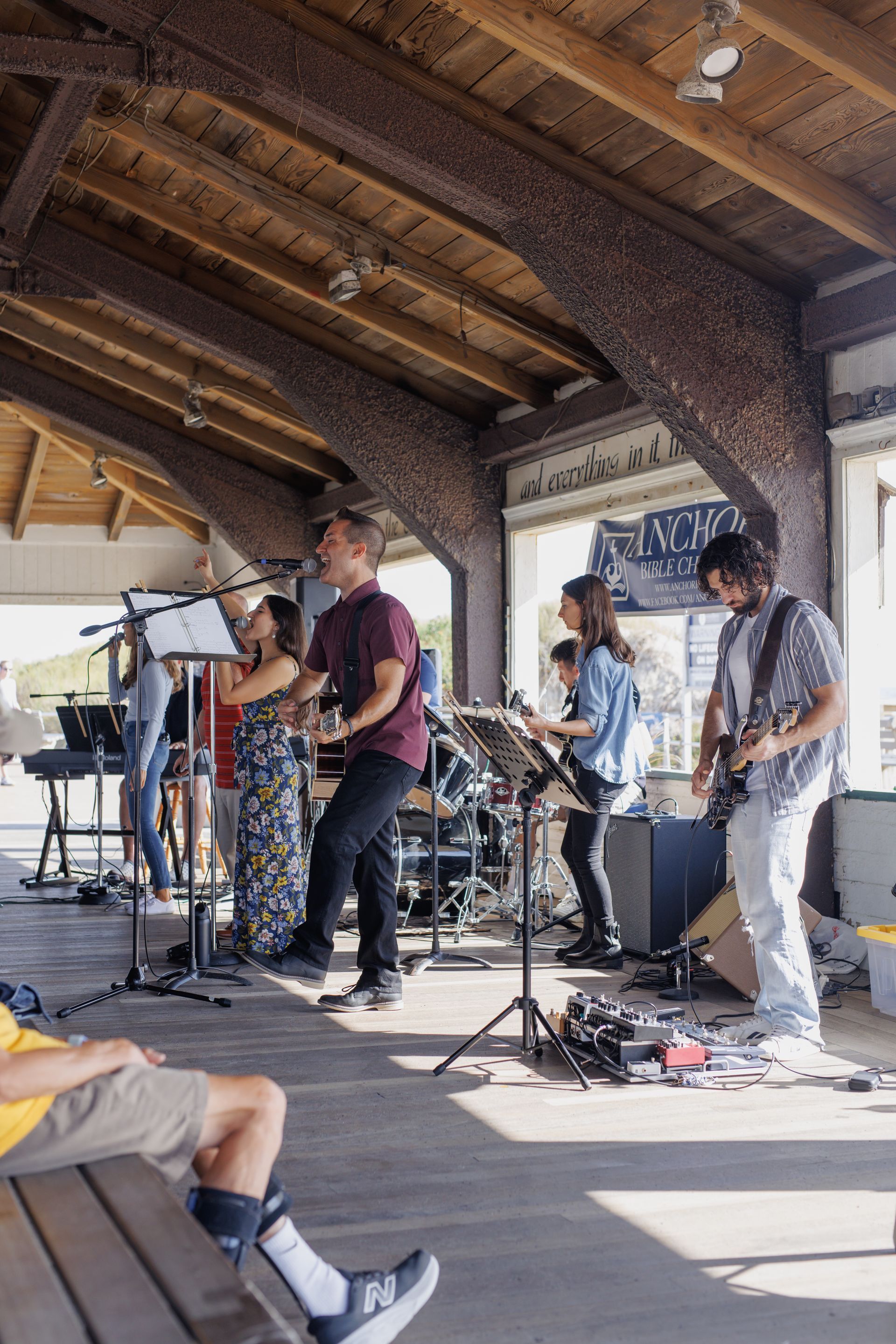 Band performing live under a wooden canopy; a woman sings, others play instruments, sunlight.
