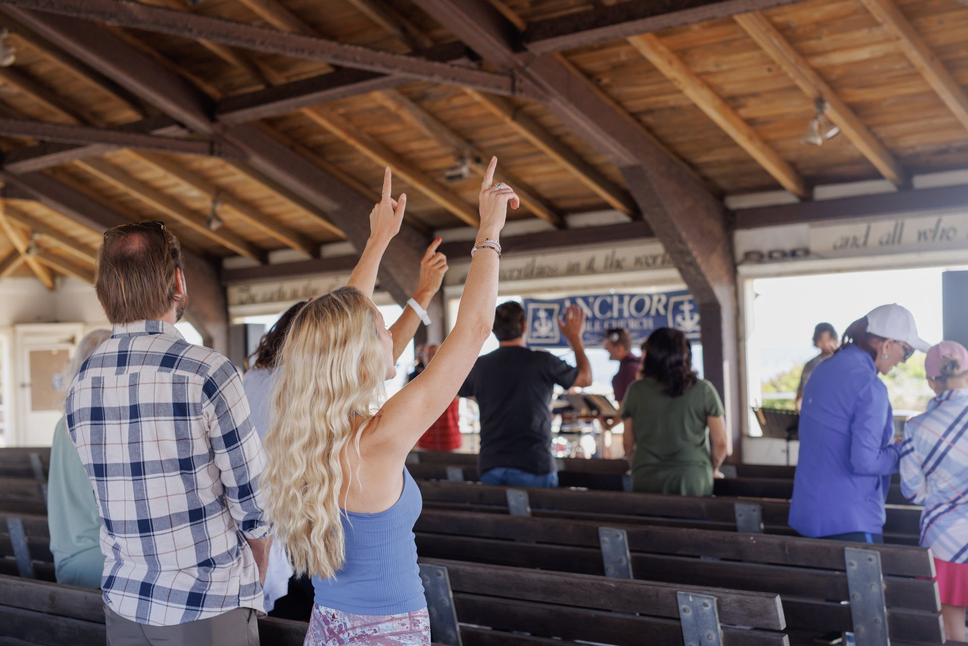 People with arms raised, in a wooden church, some facing forward, others turned away, sunny day.