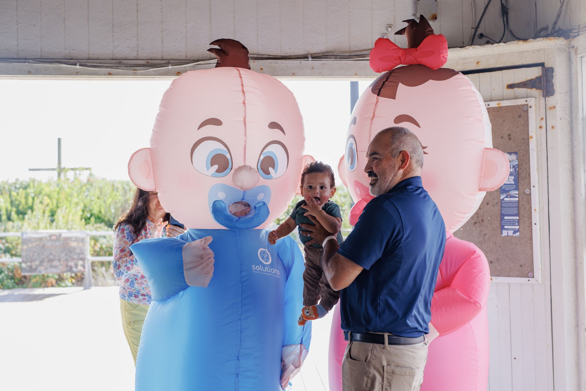 Man holds baby between large inflatable baby mascots; outdoor setting.