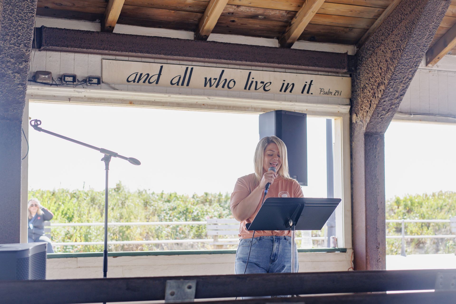 Woman speaking at a microphone outdoors. Blonde hair, casual clothes, and a wooden shelter backdrop.