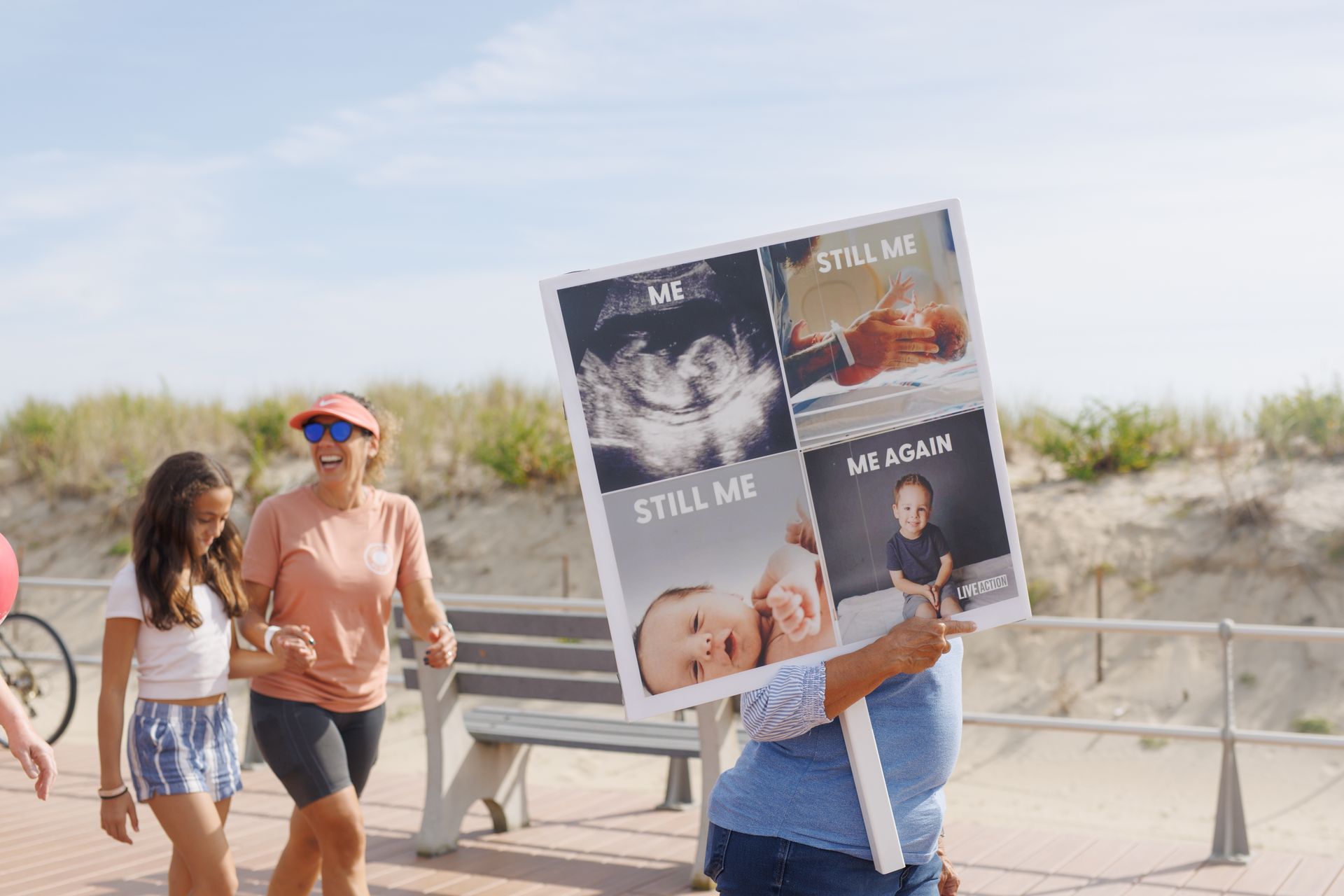 People walking on a boardwalk, one holding a sign with photos of babies, sunlit.