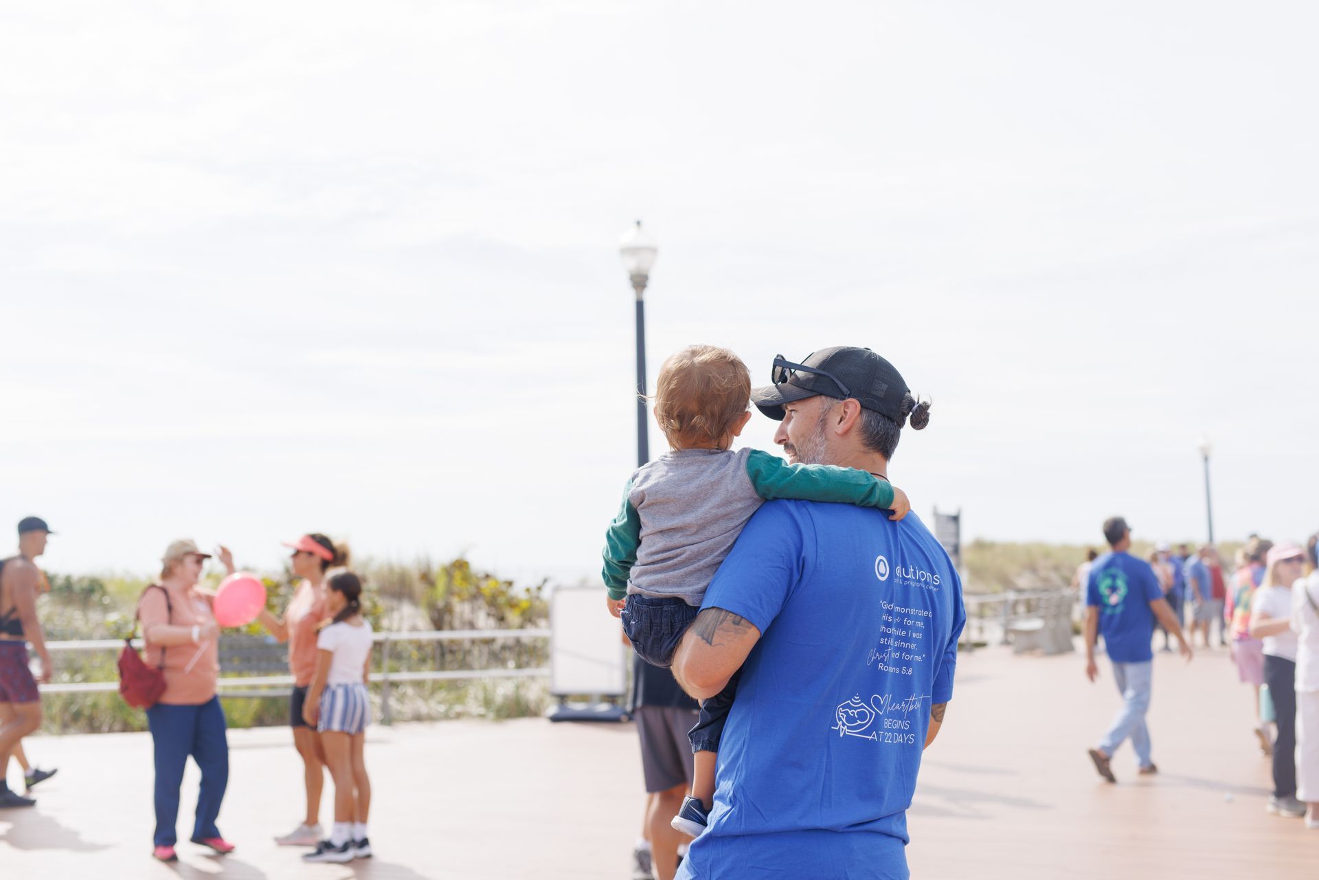Man carrying a child on a boardwalk; people walking in the background. Sunny day.