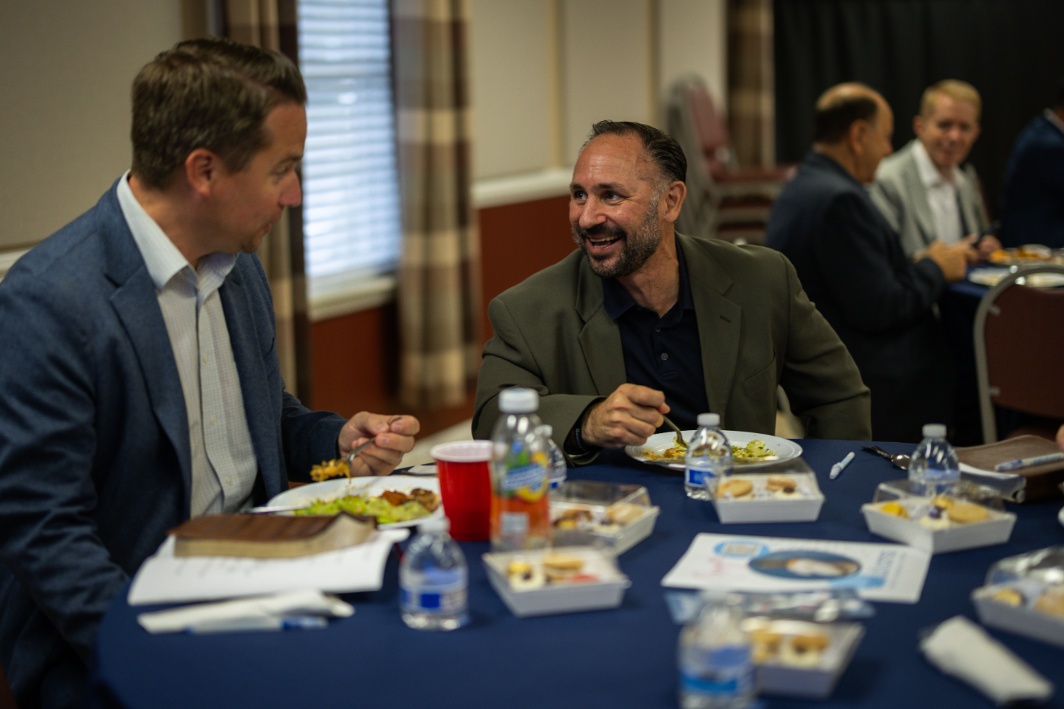 Two men are sitting at a table eating food and drinking water.
