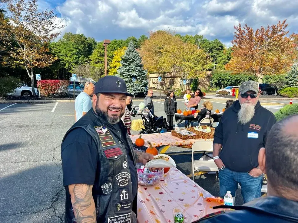 Men at an outdoor event, smiling. Table of treats and pumpkins. Fall foliage and parking lot.
