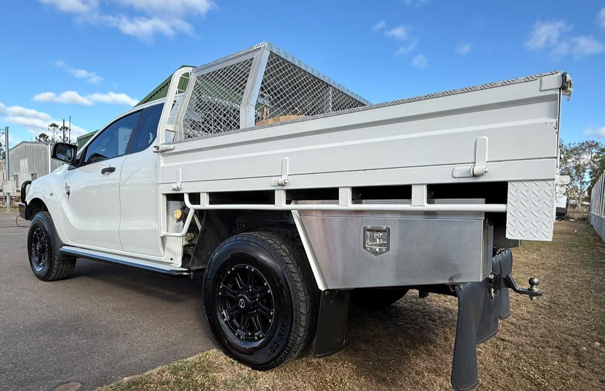 A white pickup truck with a custom tray, wire headboard, and metal tool storage box parked outdoors under a blue sky.