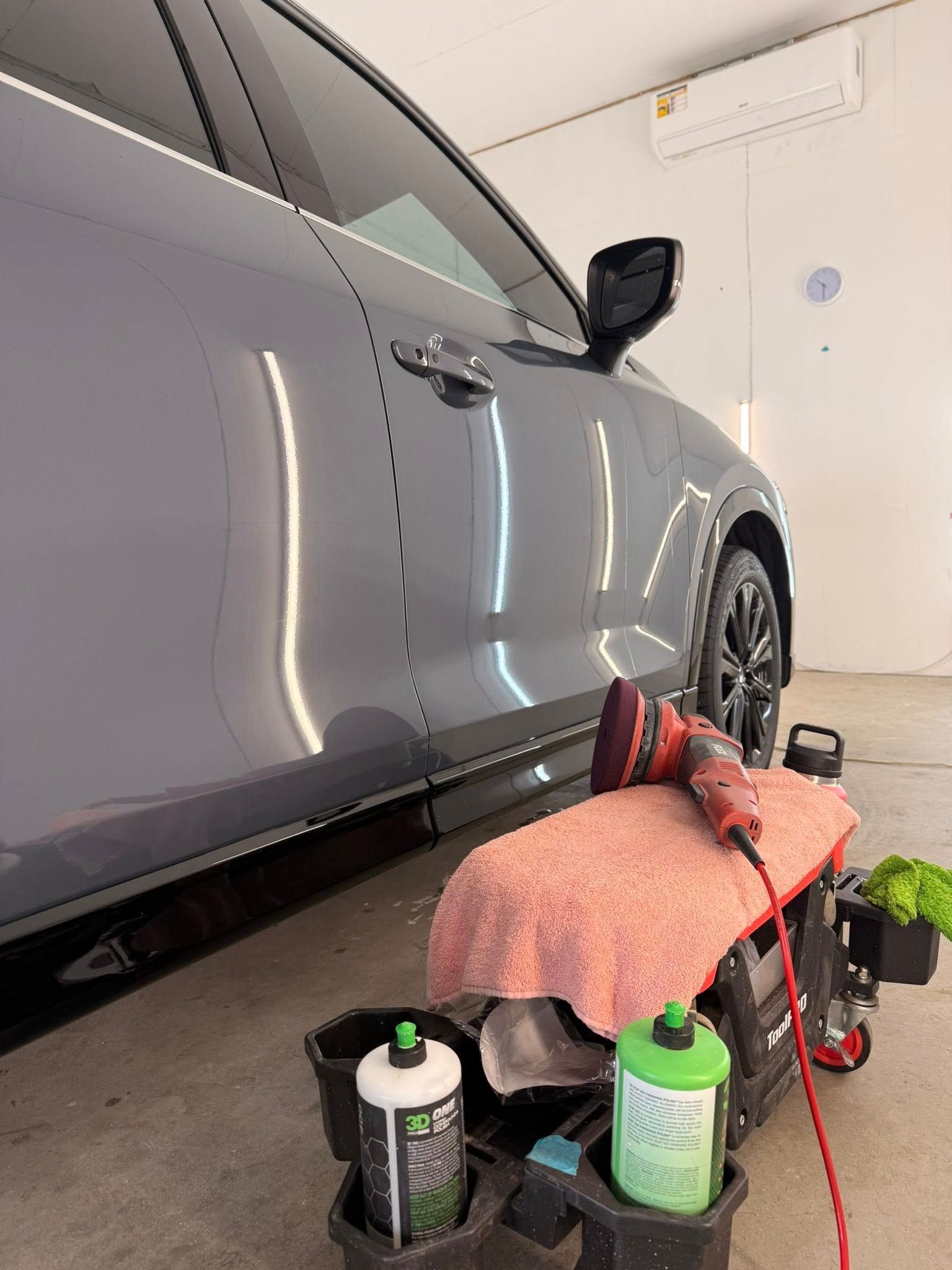 A grey car parked in a garage with a mobile detailing cart containing a polisher, cloths, and bottles of supplies.